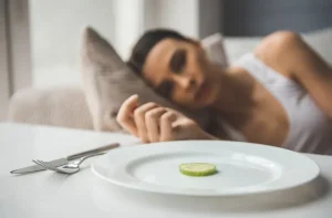 Image of a woman lying on a pillow, gazing at a plate with a single slice of cucumber on it.